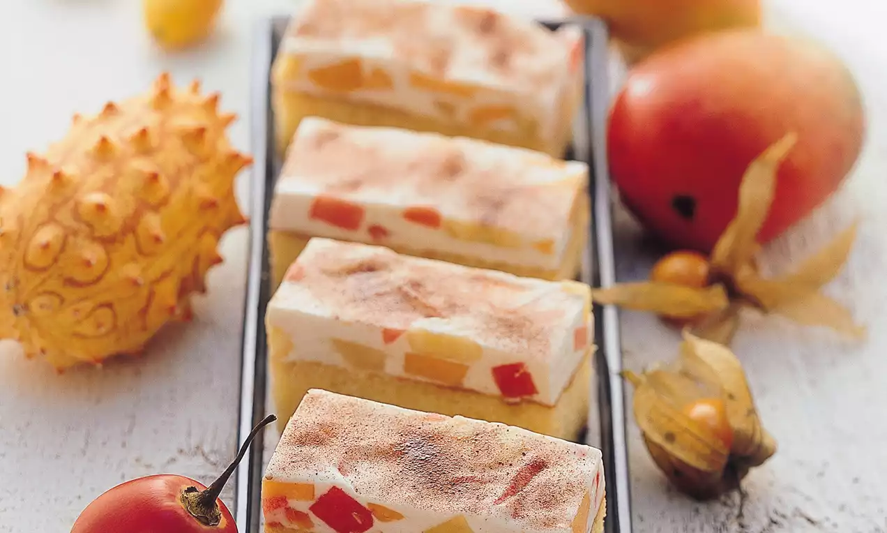 Wie rechteckige Schnitten aus hellem Biskuit mit tropischen Fruchtstücken und leichter Creme, angerichtet auf einer schmalen Platte mit exotischen Früchten