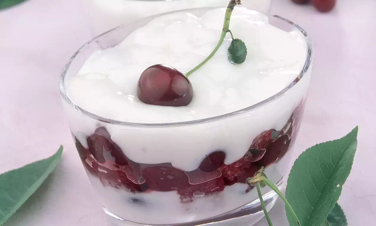 Kadeh içinde kremalı beyaz muhallebi, alt katmanında vişne parçaları, üstte taze vişne ve çevresinde yeşil yapraklarla sunulmuş