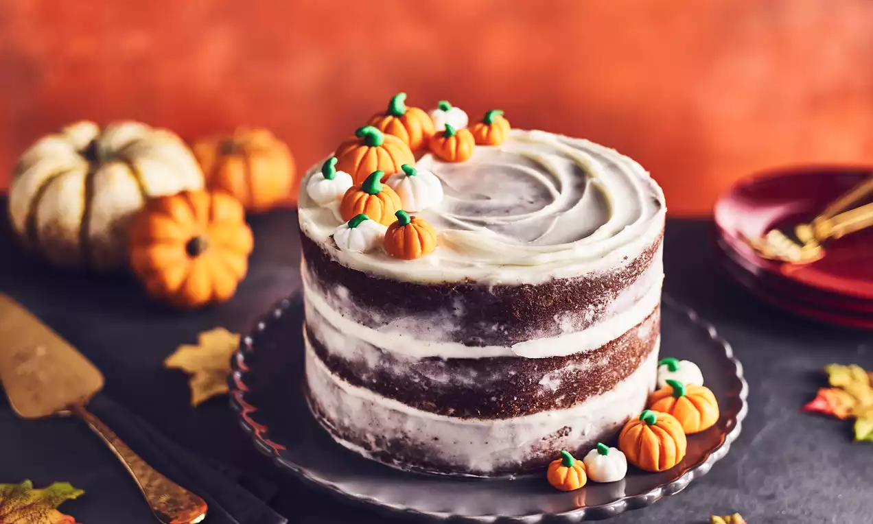Layered pumpkin cake with smooth cream frosting, topped with tiny fondant pumpkins, on a scalloped glass plate surrounded by autumn leaves