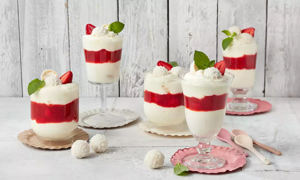 Schichtdessert in klaren Gläsern mit weißer Creme, rotem Erdbeerpüree, Sahnehäubchen, Raffaello-Kugel und Minzblatt auf pastellfarbenen Tellern