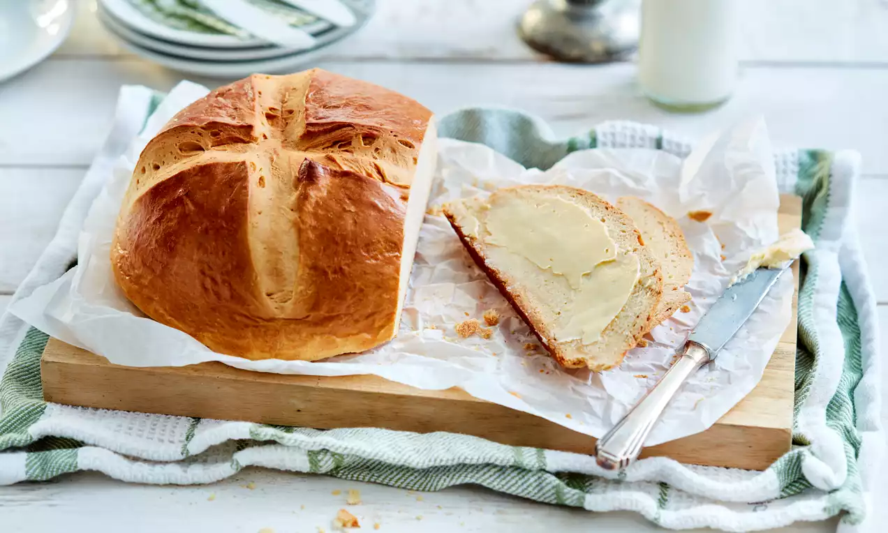 Goldbraunes Hefe-Osterbrot mit Kreuzschnitt, ein dick bestrichenes Stück daneben, auf Backpapier und Holzbrett mit Messer angerichtet