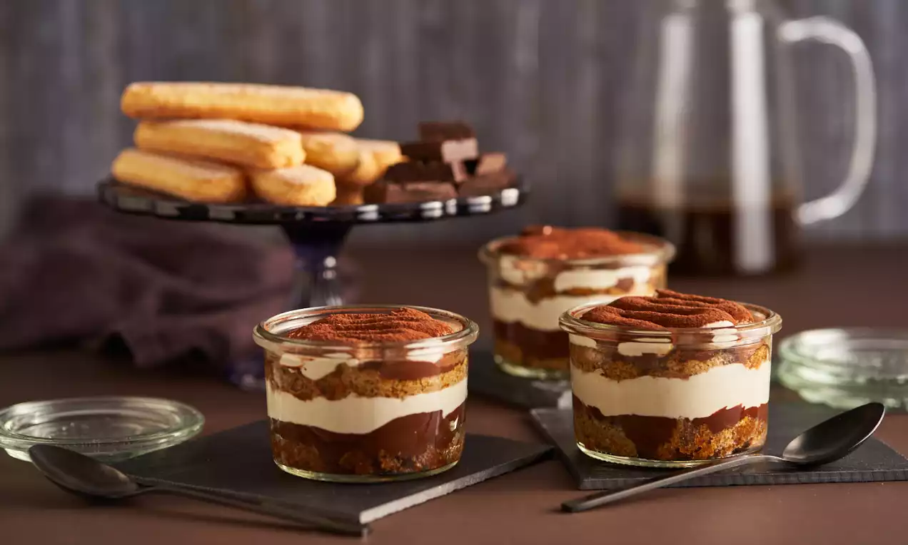 Piccoli bicchieri di vetro con strati alternati di crema chiara al mascarpone, biscotto marrone, cacao spolverato e cioccolato fondente