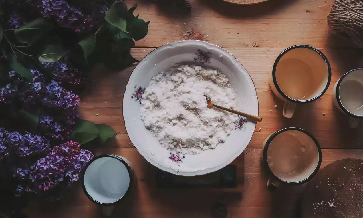 Bílý prášek jemné struktury v květovaném porcelánovém talíři s dřevěnou lžící na dřevěném stole obklopeném hrnky a větvemi šeříku