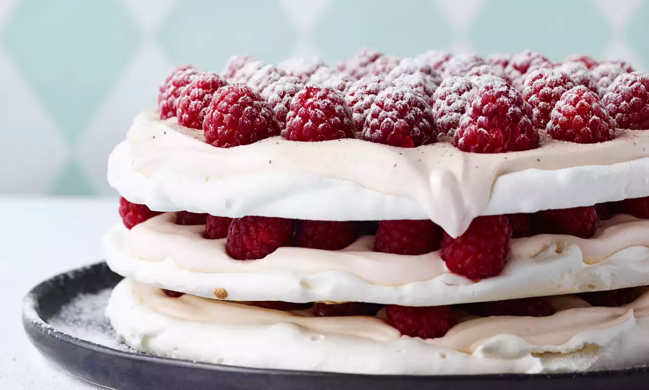 Kolmikerroksinen valkoinen marenkikakku pehmeällä vaaleanpunaisella täytteellä ja punaisilla vadelmilla, tomusokerilla ripoteltuna tummalla lautasella