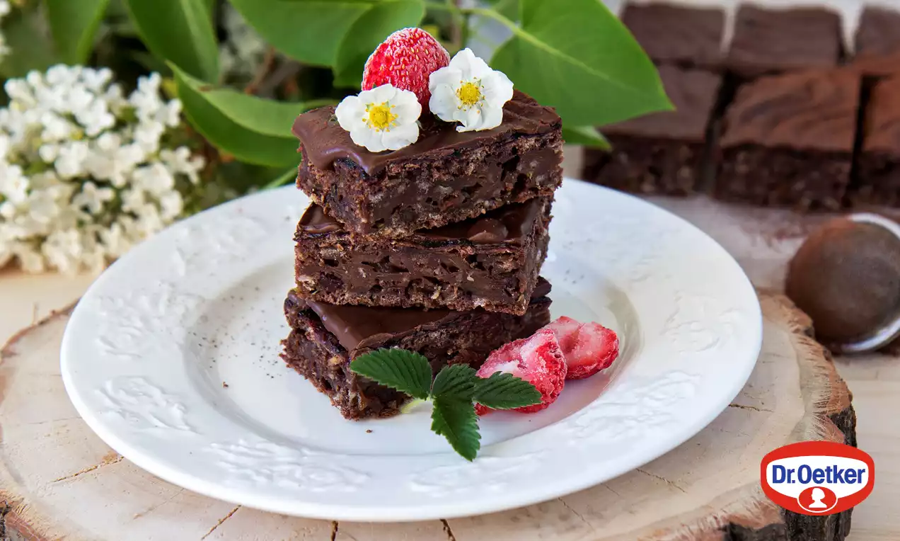 Tmavohnedé čokoládové kocky s lesklou polevou poukladané na seba, ozdobené jahodou, bielymi kvietkami a lístkom na bielom tanieri