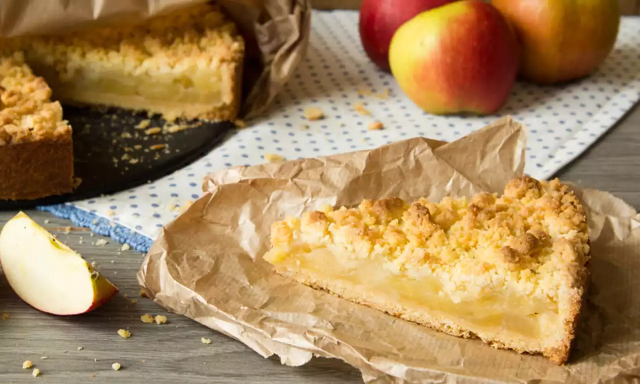 Part de tarte aux pommes dorée avec une croûte épaisse et un crumble croustillant, posée sur du papier kraft avec des pommes en arrière-plan