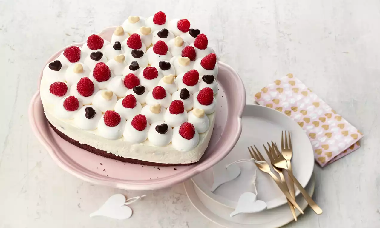 Srcoliki kolač s čokoladnom podlogom, kremastim bijelim slojem, ukrašen svježim malinama, čokoladnim i bijelim srcolikim dekoracijama na ružičastom tanjuru