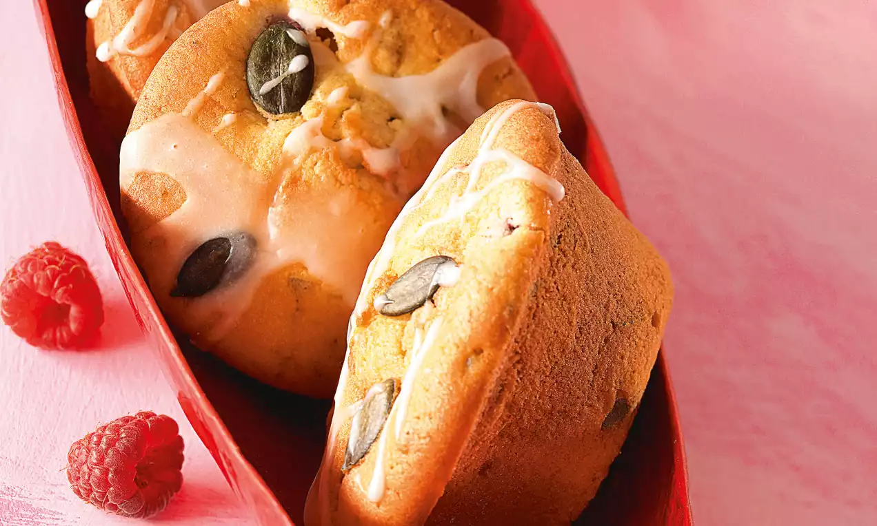 Muffin soffici dorati con semi di zucca e glassa bianca lucida, disposti in un vassoio rosso su uno sfondo rosa pastello con lamponi freschi