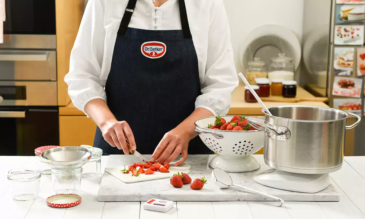 Eine Person bereitet auf einer weißen Arbeitsfläche frische rote Erdbeeren vor, neben einem Sieb und einem Topf in einer Küchenumgebung