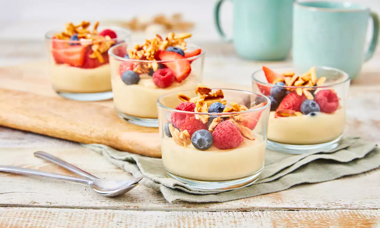 Svijetložuta kremasta mandel puding baza u staklenim čašama, ukrašena svježim jagodama, malinama, borovnicama i smeđim granolom