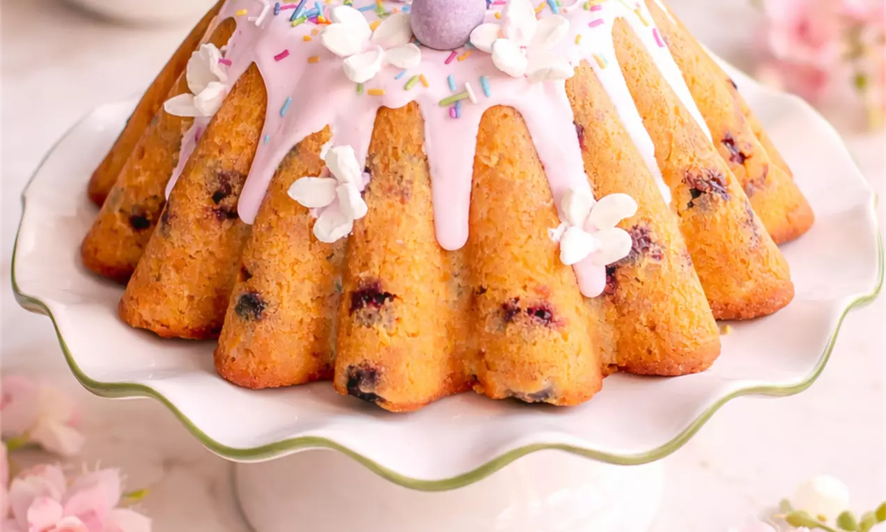 Zlatnosmeđi kuglof s tamnim voćnim komadićima, preliven svijetloružičastom glazurom, šarenim posipom i ukrasnim jajima na bijelom postolju