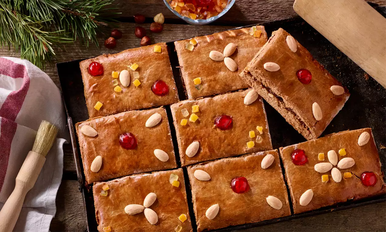 Rechtecke aus glänzendem braunem Lebkuchenteig, verziert mit roten kandierten Kirschen, Mandeln und gelben Zuckerstückchen auf einem dunklen Blech