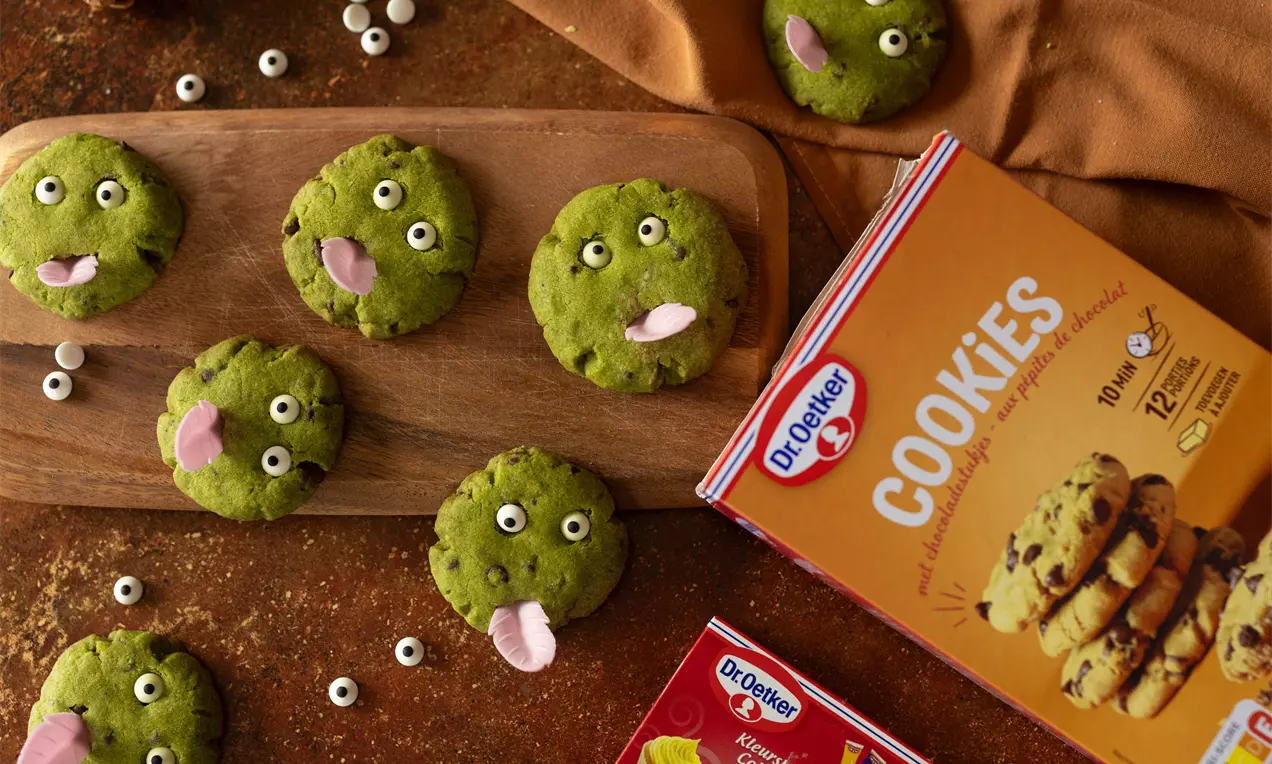 Groene ronde koekjes met ogen en roze tongen, speels gerangschikt op een houten plank en bruine ondergrond met Dr. Oetker-verpakking