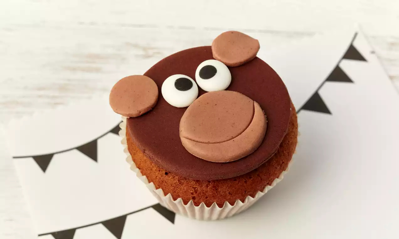 Ein Muffin mit braunem Fondant als Affengesicht, hellbraunen Ohren, Schnauze und cremeweißen Augen auf weißem Papier mit Dreiecksmuster