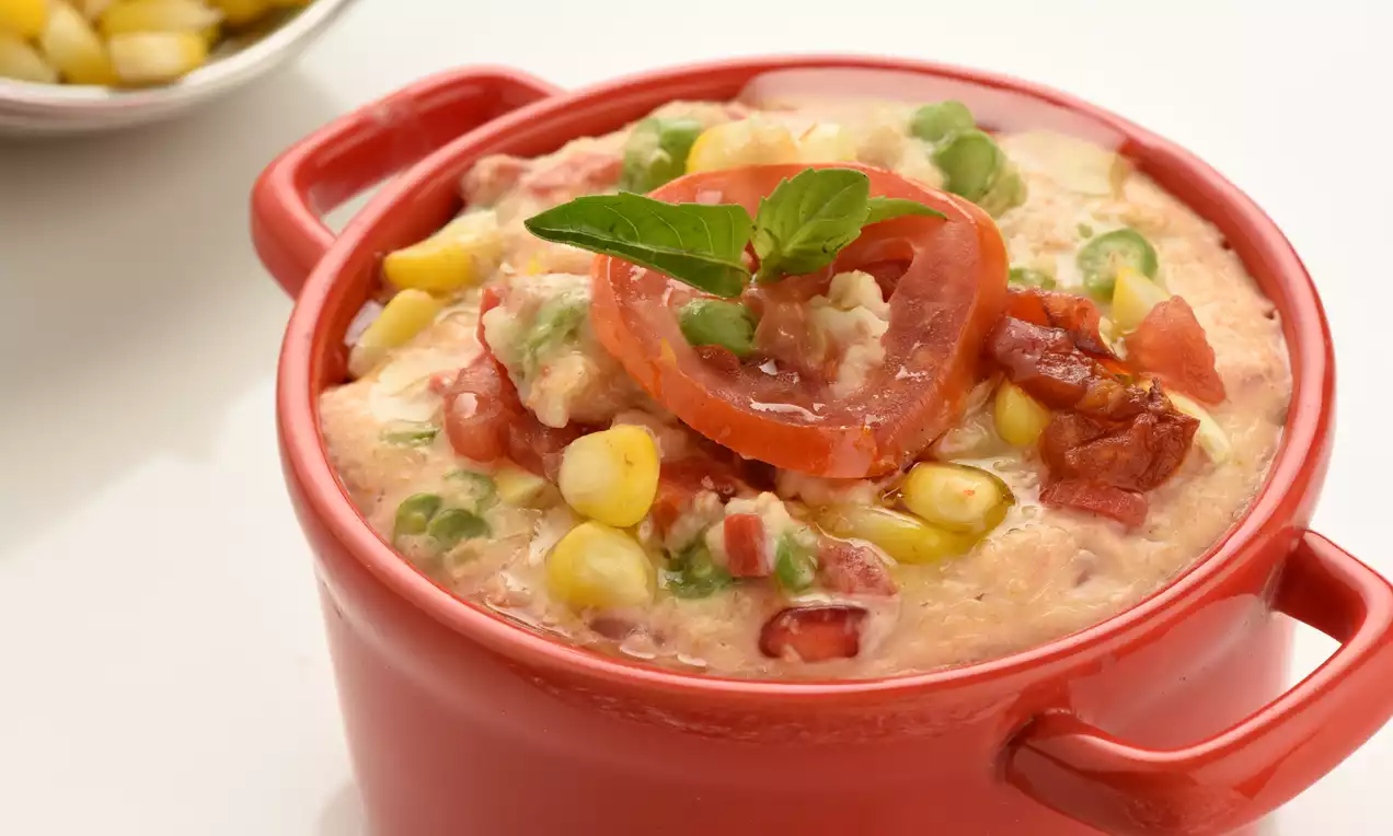 Creamy oatmeal with a pinkish hue in a red pot, topped with a slice of tomato, green peas, yellow corn, crispy bacon and a basil leaf 