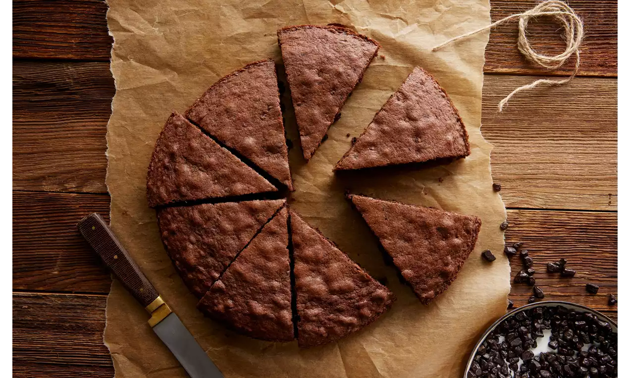 Yumuşak dokulu koyu renkli çikolatalı turta, dilimlenmiş şekilde kahverengi pişirme kağıdı üzerinde, yanında çikolata parçaları ve bir bıçak ile yerleştirilmiş