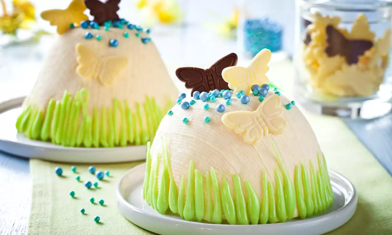 Waniliowa pascha w jasnym kremowym kolorze o zaokrąglonym kształcie, dekorowana zielonym kremem i czekoladowymi motylami na białym talerzu