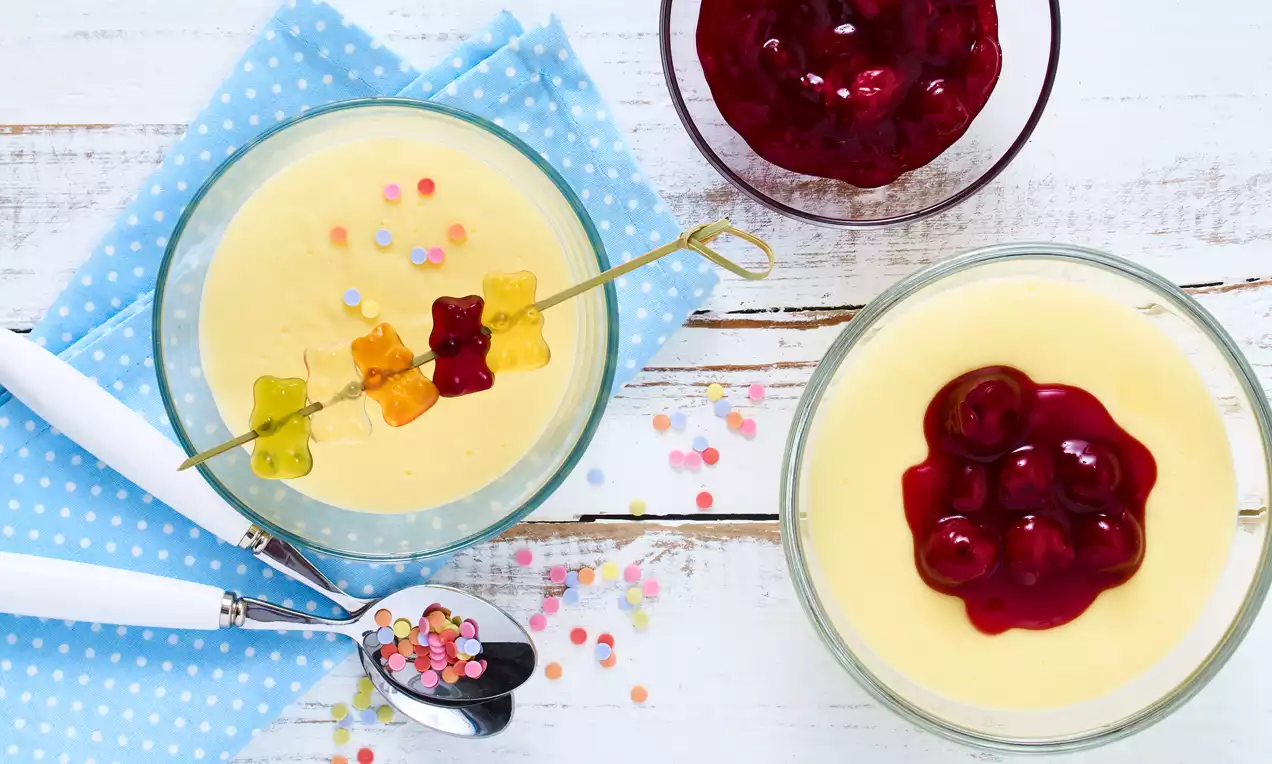 Heller Vanillepudding in Glasschalen mit Gummi-Bärchen-Spieß und Kirschsoße, dekoriert mit bunten Zuckerstreuseln auf gepunktetem Stoff