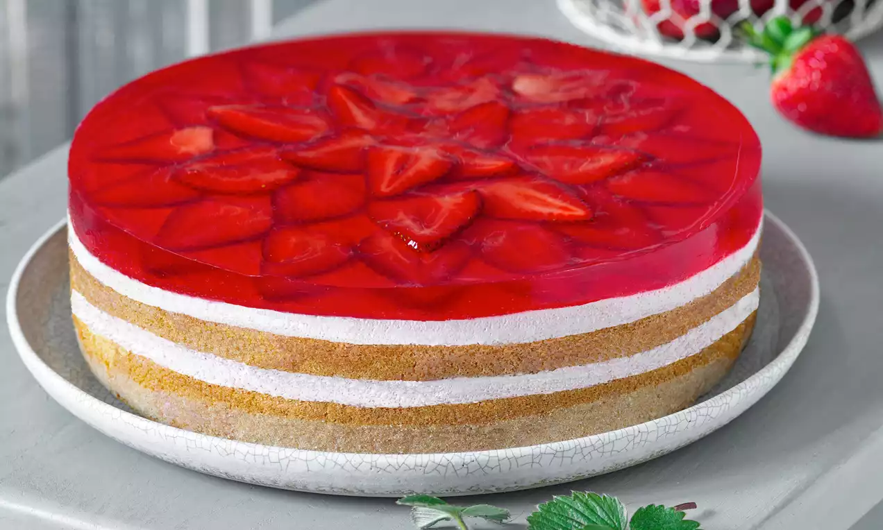 Jahodovo smotanový dort s vrstevnatým základom zlatistého korpusu a bielej náplne, hladkým červeným želé a plátkami jahôd na tanieri