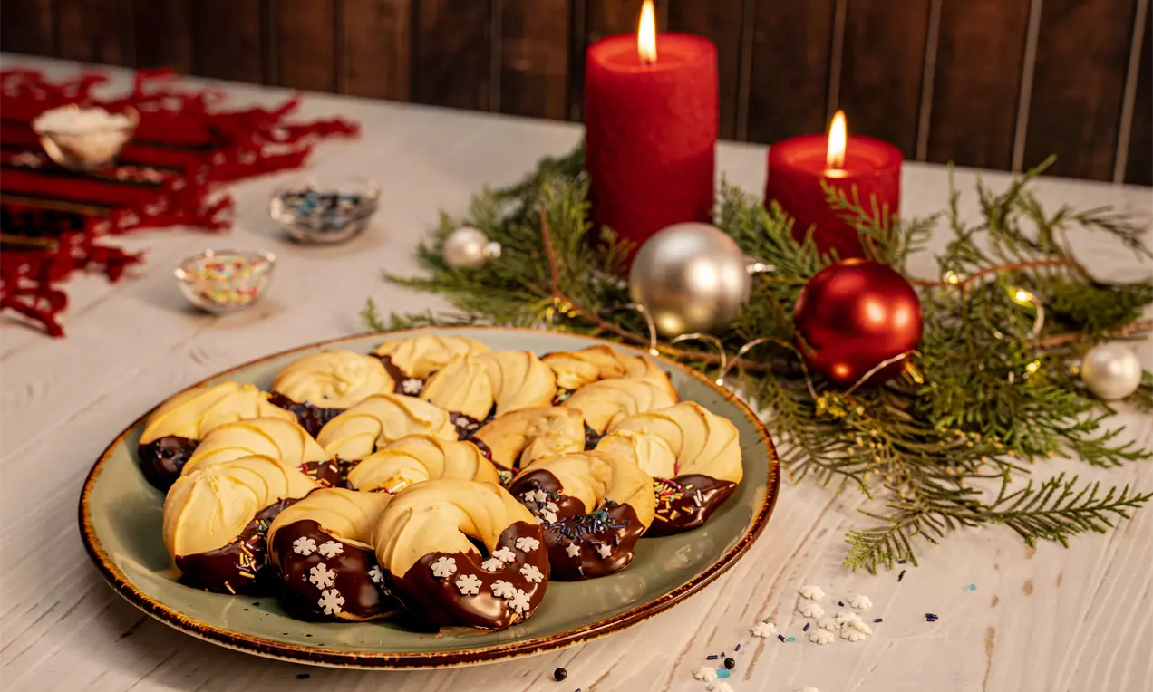 Fursecuri aurii sub formă de semilune, glazurate parțial cu ciocolată lucioasă și ornamente delicate, aranjate pe o farfurie verde lângă decorațiuni de Crăciun