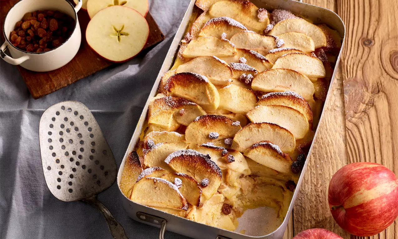 Goldbrauner Brotpudding mit weichen Apfelscheiben, Rosinen und Puderzucker in einer rechteckigen Form auf rustikalem Holzbrett