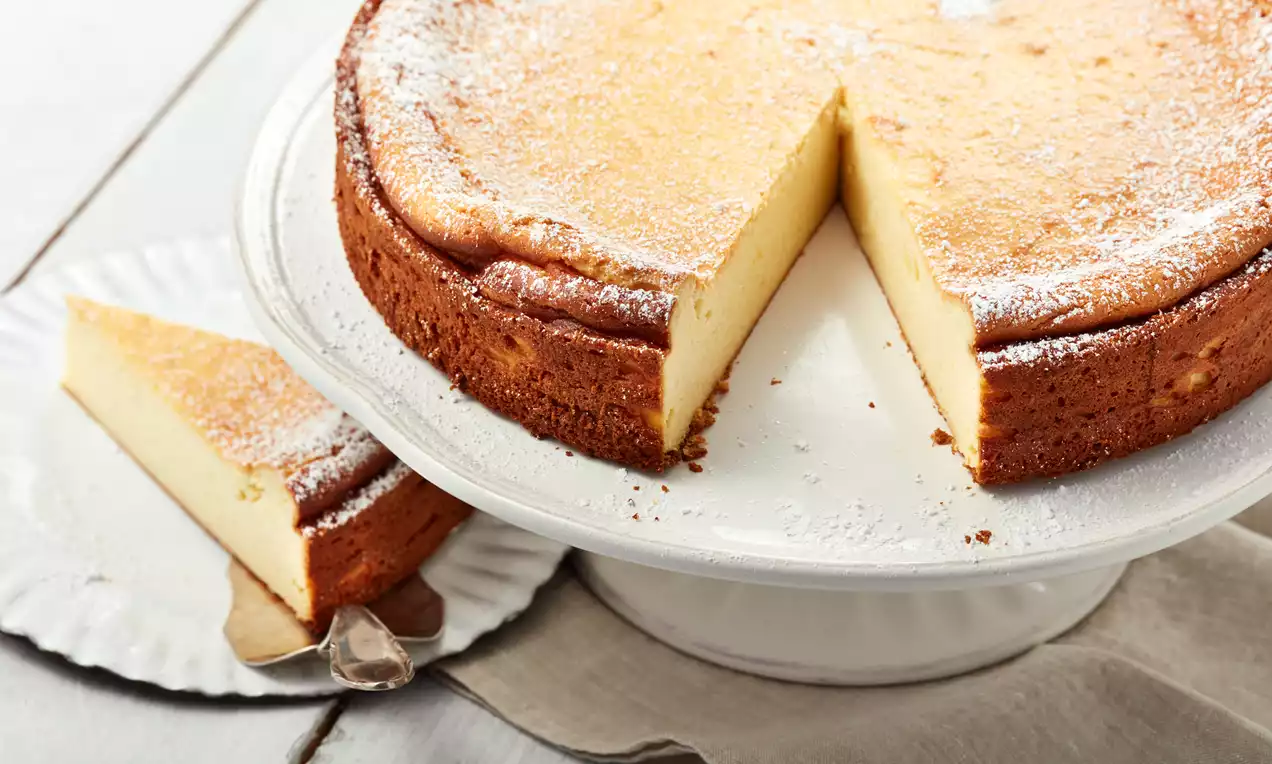 Goldbrauner Käsekuchen ohne Boden mit leichter Puderzuckerhaube auf weißer Tortenplatte, angeschnitten, ein Stück auf silberfarbenem Tortenheber