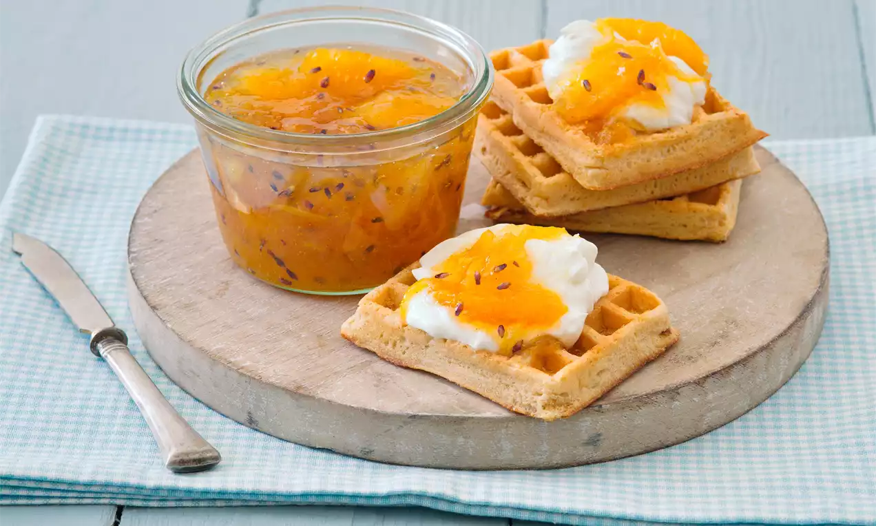 Citrusová marmeláda jemne oranžovej farby v sklenenej nádobe vedľa zlatistých vaflí s kopčekom smotany a plátkami pomaranča na drevenej podložke