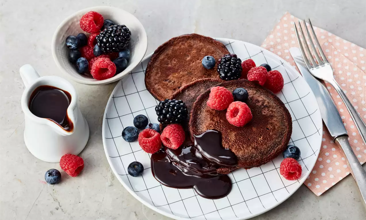 Zwei dunkelbraune Schoko-Pancakes mit glänzender Schokosauce, frischen roten Himbeeren, Brombeeren und Blaubeeren auf einem weißen Teller mit Gittermuster