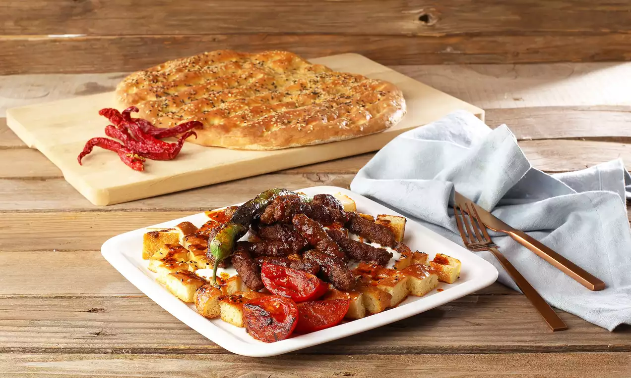 Küp şeklinde kızarmış pide üzerinde soslu köfteler, közlenmiş biber ve domateslerle beyaz tabakta sunulmuş, yanında taze pide ve kurutulmuş biberler