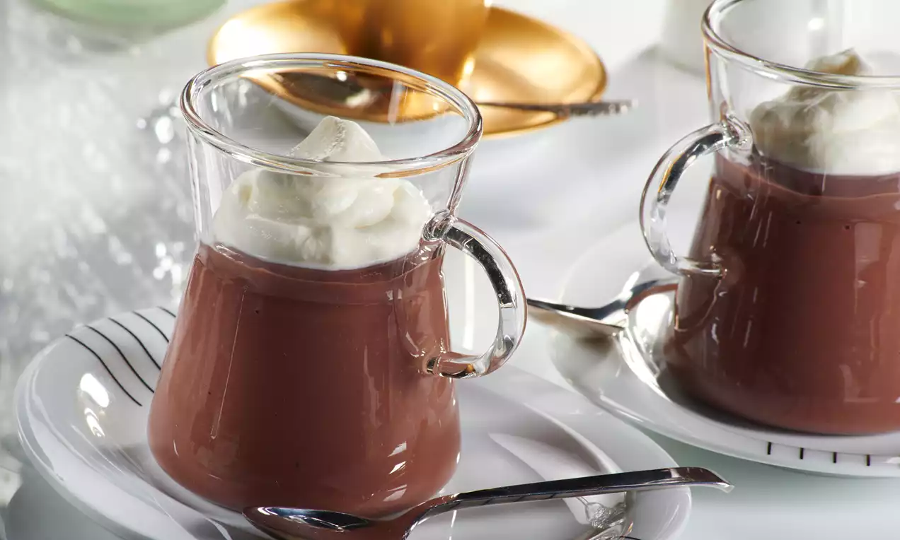 Pudim de chocolate marrom brilhante servido em xícara transparente com camada suave de chantilly branco no topo sobre prato com colher prateada