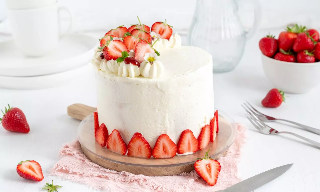 Visoka torta prekrivena bijelom kremom, ukrašena svježim jagodama na vrhu i slojem tankih kriški jagoda uz donji rub, na drvenom podlošku