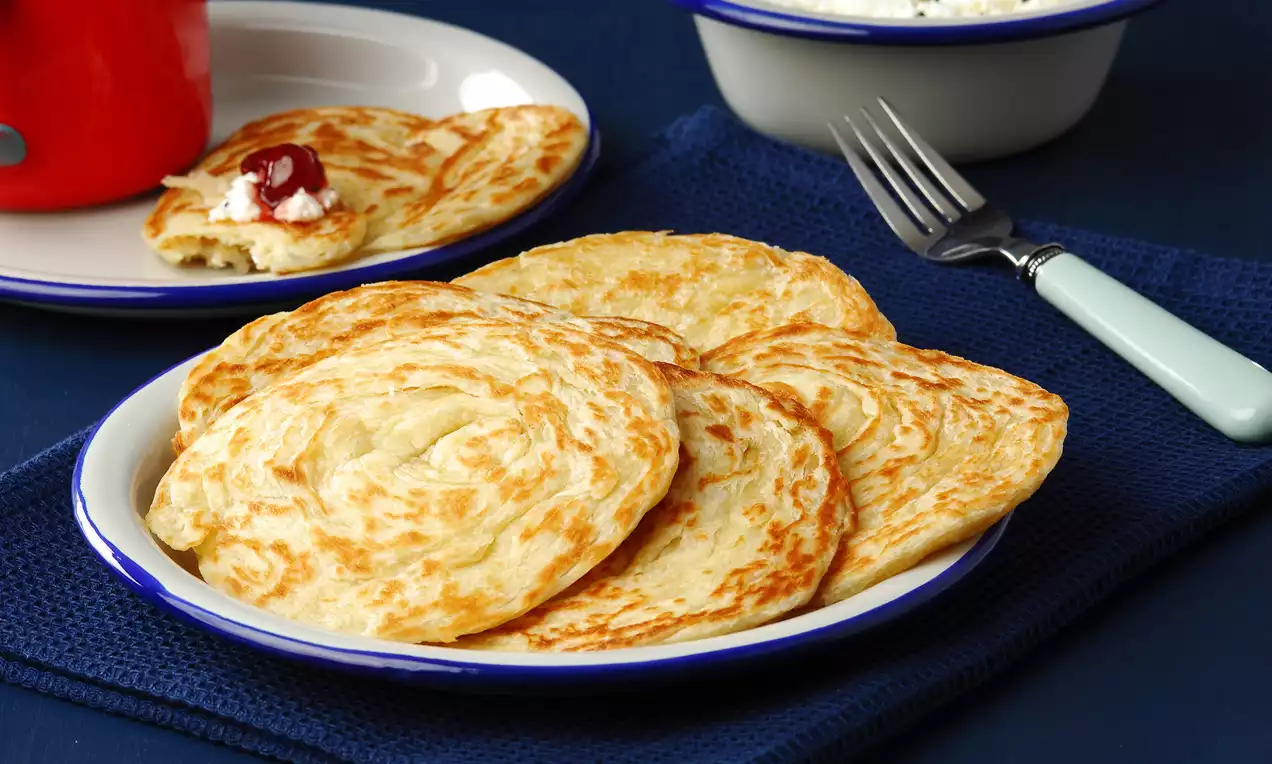 Altın rengi, ince katmanlar halinde kıvrımlı tereyağlı katmerler, beyaz kenarlı tabakta mavi bir örtü üzerinde düzenlenmiş