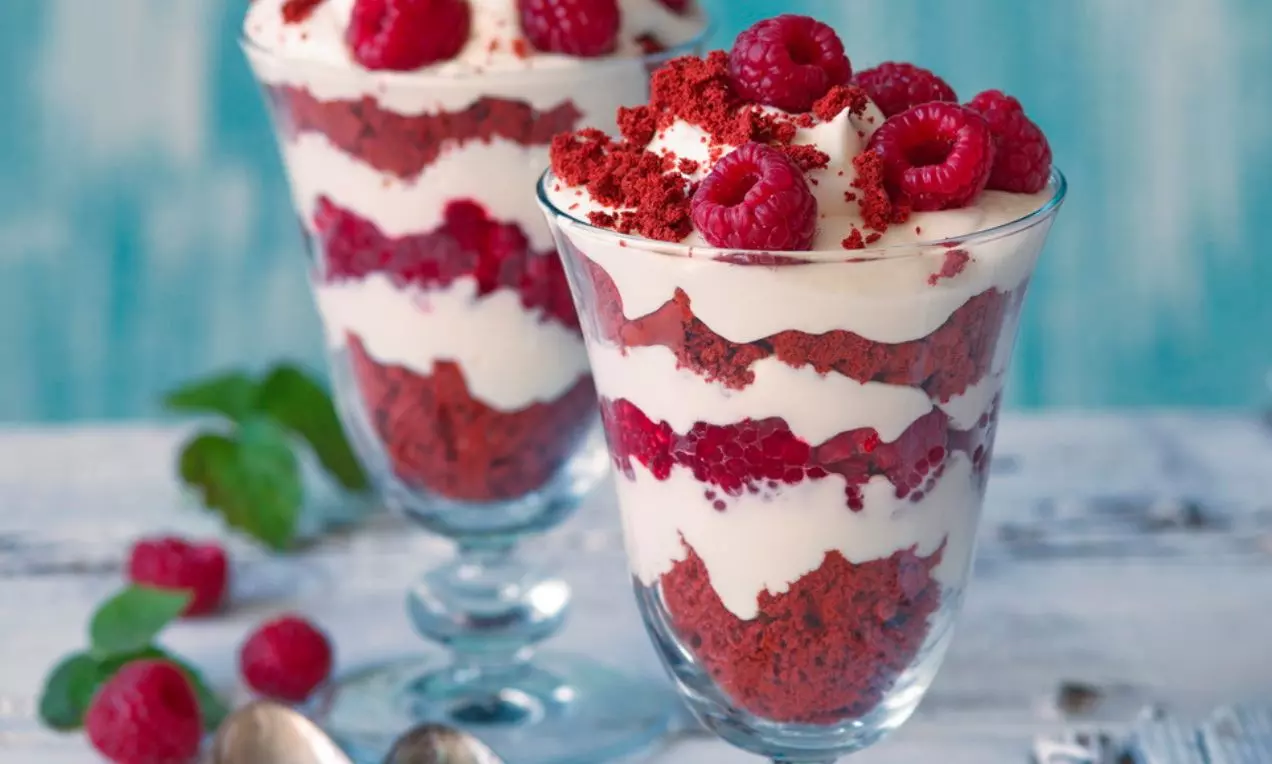 Svetlý sklenený pohár s vrstvami červeného red velvet cesta, jemného bieleho krému, ozdobený čerstvými malinami na rustikálnom stole