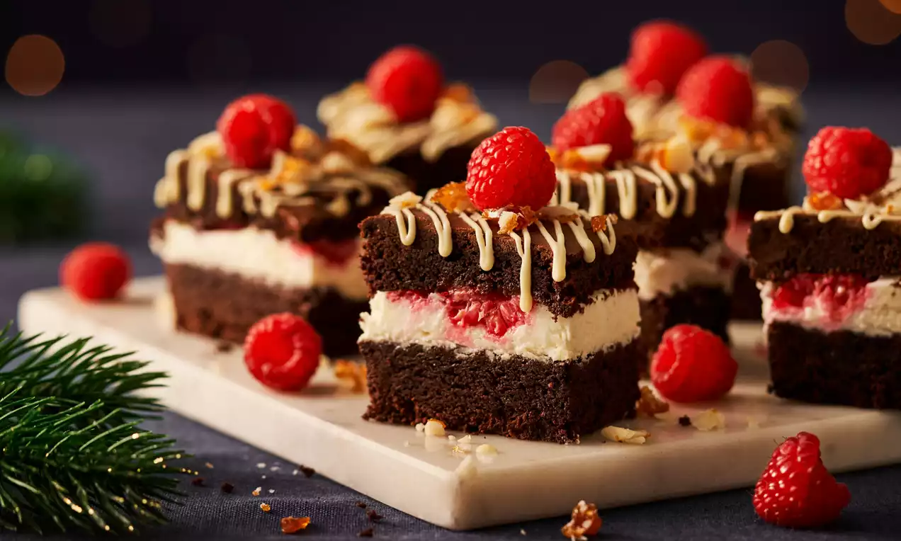 Brownie carré en couches brun foncé avec crème blanche et framboises rouges, décoré de chocolat blanc et amandes sur un plateau marbré