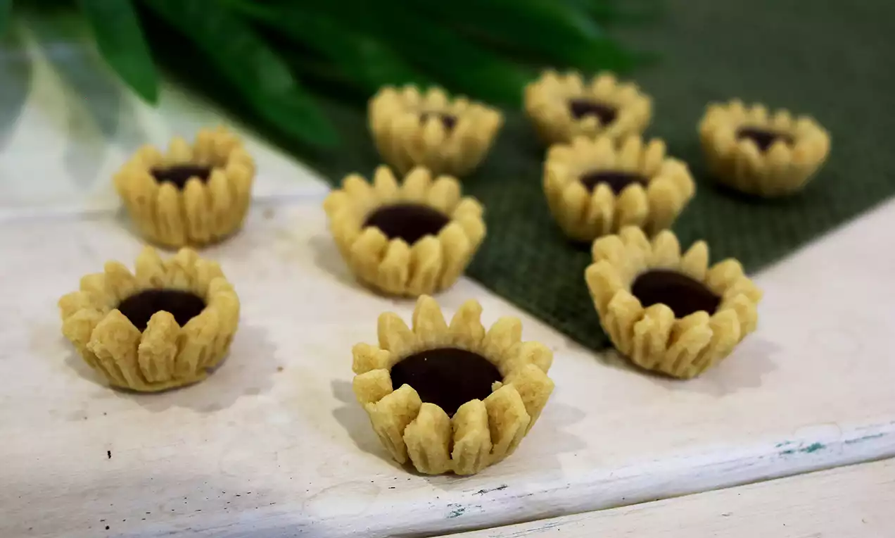 Sunflower-shaped tarts with golden, textured pastry petals and glossy dark chocolate centers arranged on a white surface with a green mat