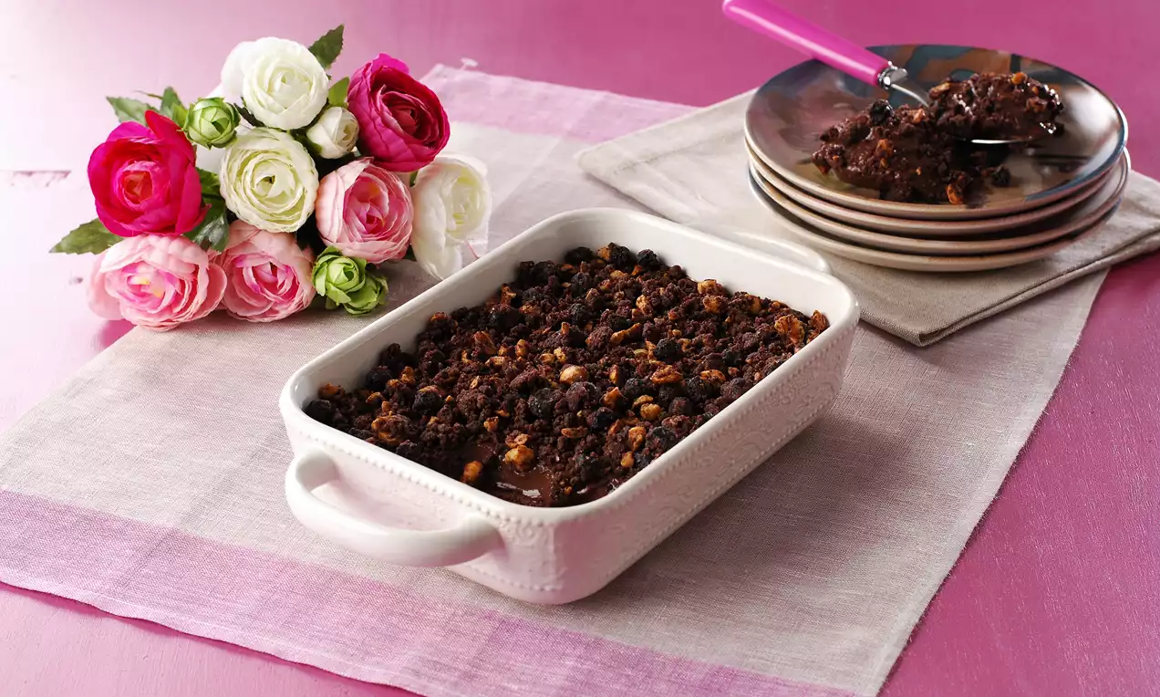Beyaz seramik bir kabın içinde koyu kahverengi kıtır crumble, pembe bir masa örtüsünde, yanında renkli çiçekler ve tabaklar ile sunulmuş