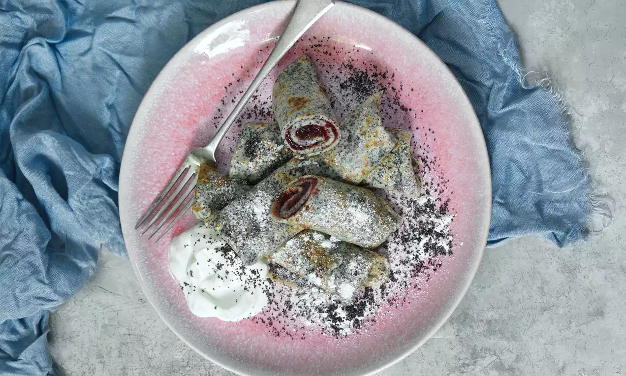 Na světlém talíři položeném na šedém povrchu jsou tenké palačinky s tmavými makovými tečkami, dekorované modrou látkou v pozadí