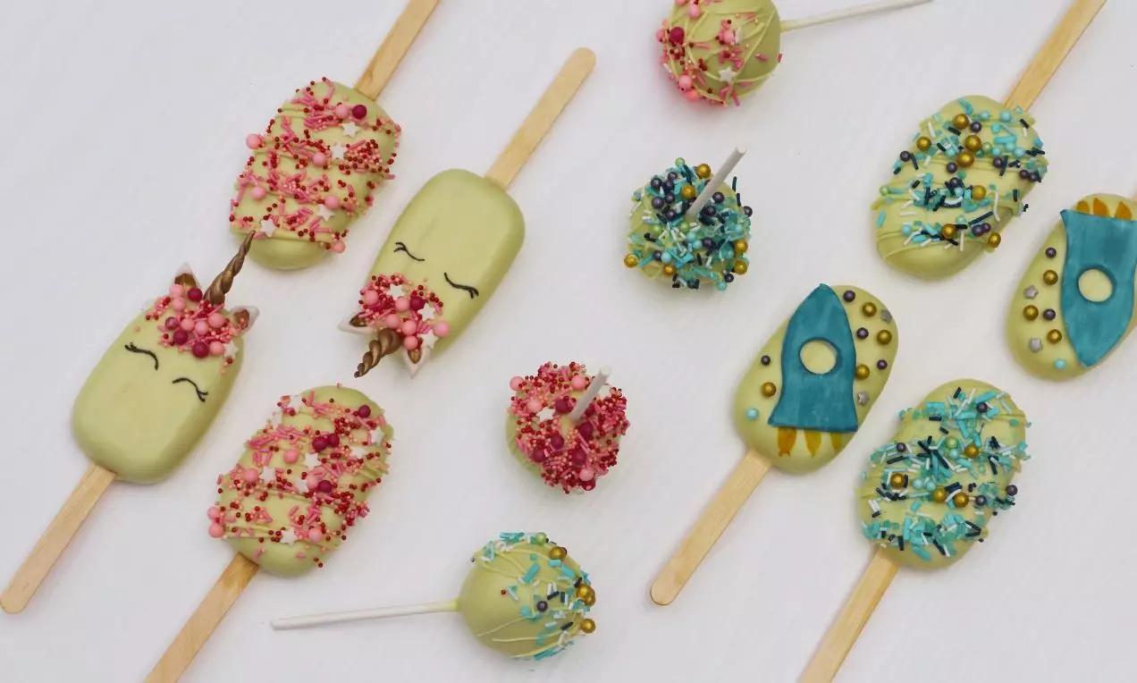 Banánově zelené nanuky na světlých dřevěných tyčinkách zdobené růžovými, modrými a zlatými detaily vedle kulatých cakepops na bílém podkladu