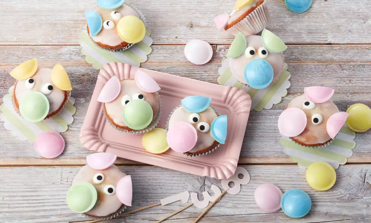 Glücksschweinchen aus rosafarbenem Zuckerguss mit bunten Ohren und runden Nasen auf pastellfarbenem Tablett und Holzuntergrund liegend