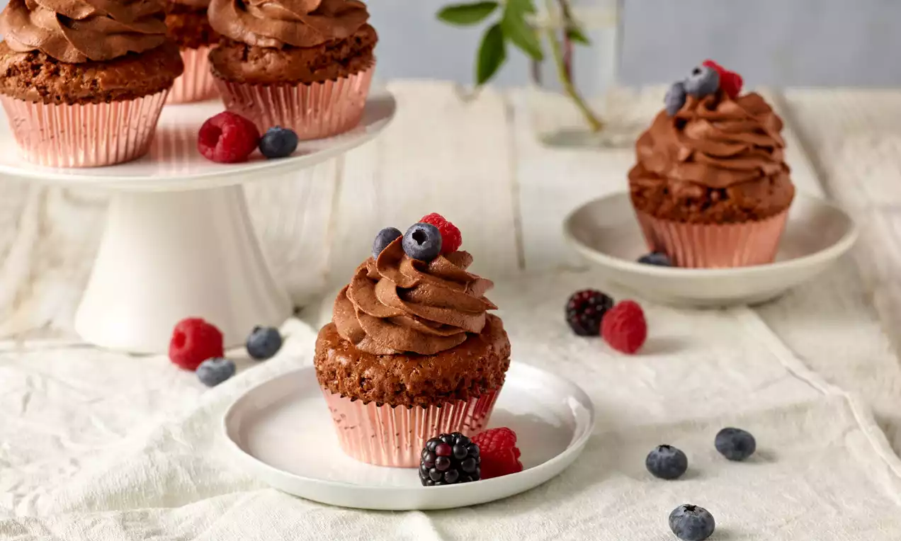 Vegane Kaffee-Schoko-Cupcakes mit fluffigem Schokofrosting in roségoldenen Förmchen, dekoriert mit frischen Beeren auf Tellern und Tortenständer
