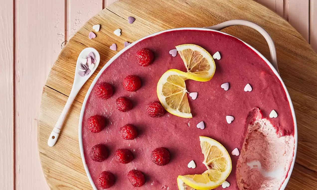 Ahududu ve limon dilimleriyle süslenmiş kırmızı kremalı tatlı, yuvarlak beyaz tabakta ahşap sunum zemininde yerleştirilmiş