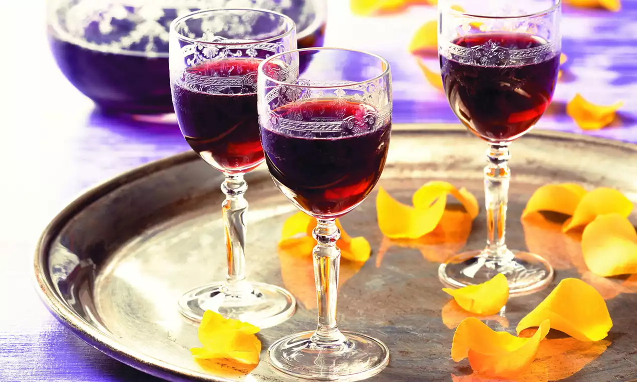 Dunkelroter Holunder-Likör in eleganten, gläsernen Kelchgläsern auf einem silbernen Tablett, umgeben von gelben Blütenblättern