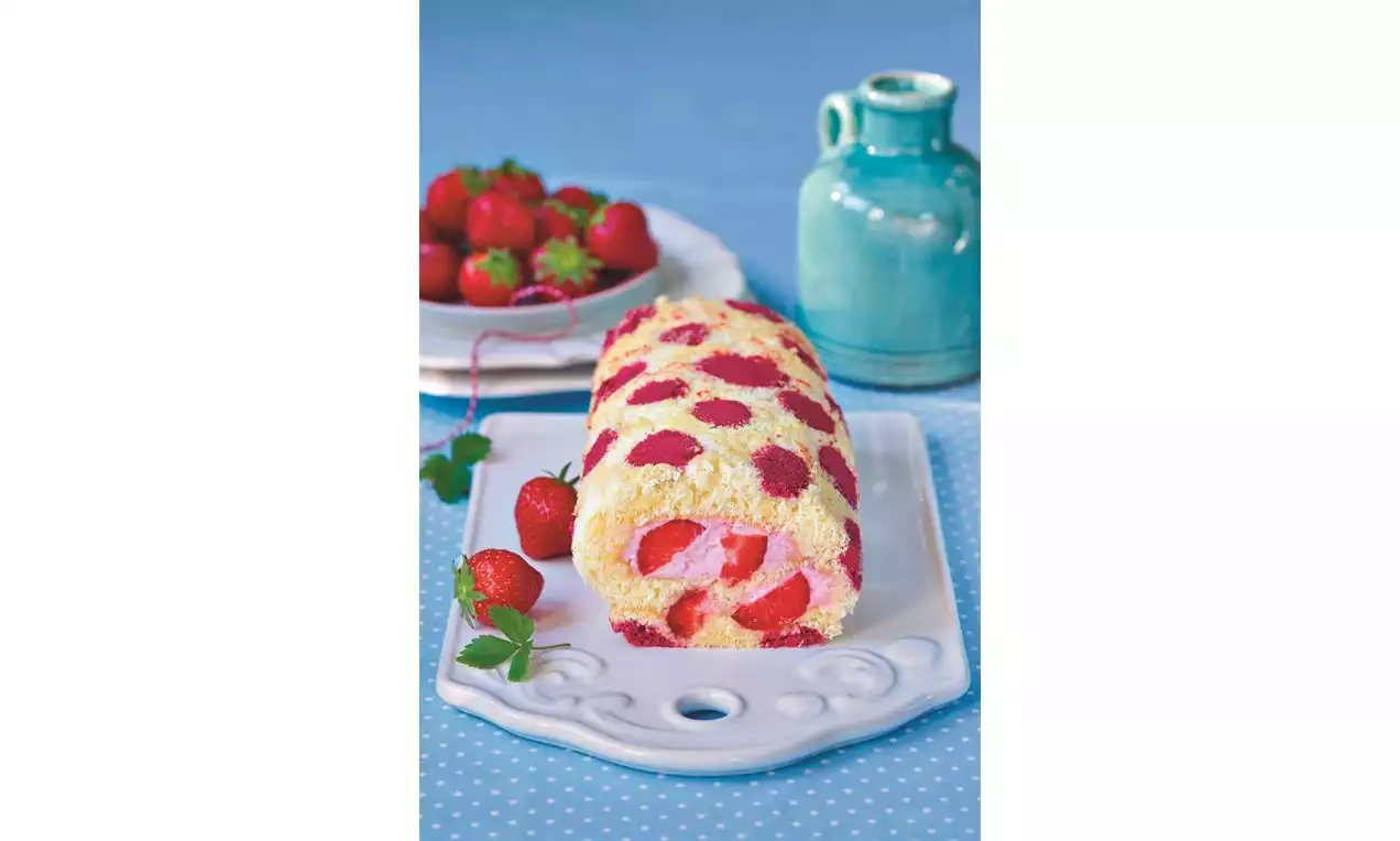 Světlá piškotová roláda zdobená červenými puntíky, uvnitř jahodová náplň, lehce položená na bílém talíři s čerstvými jahodami vedle