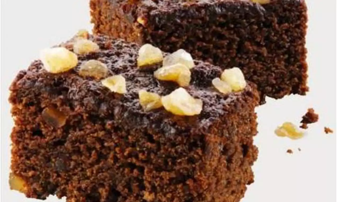 Two square pieces of dark brown gingerbread with a moist texture topped with glossy golden chunks of candied ginger on a white surface