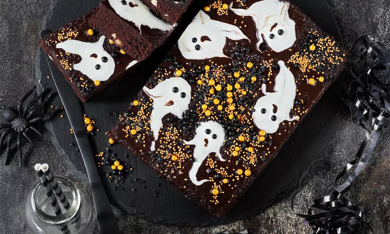 Čtvercové čokoládové brownies na černém talíři, zdobené bílými cukrovými duchy, žlutými a oranžovými cukrovými perličkami, s temnou výzdobou okolo