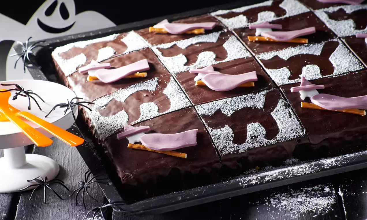Schwarzer Blechkuchen mit glänzender Schokoglasur, quadratisch geschnitten, verziert mit Puderzucker-Motiven, Hexenbesen und lila Deko