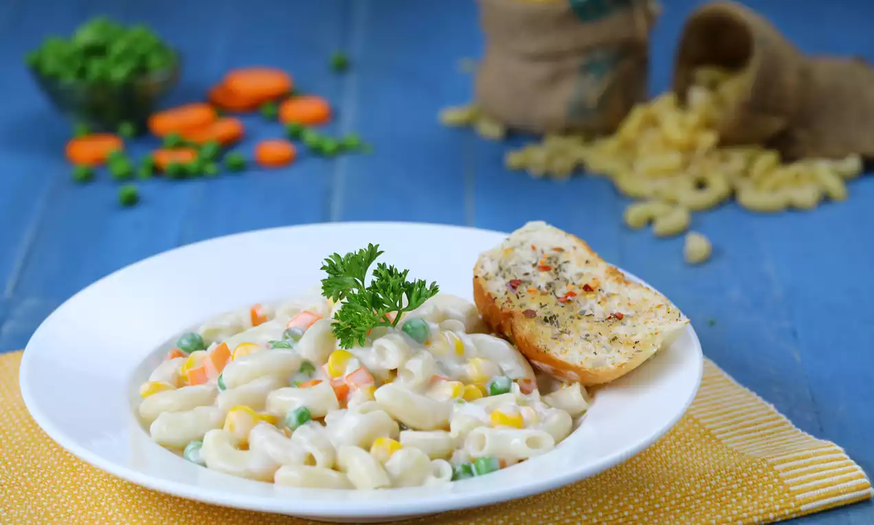 Elbow macaroni coated in creamy white sauce mixed with peas, carrots, and corn, garnished with parsley, served with garlic bread