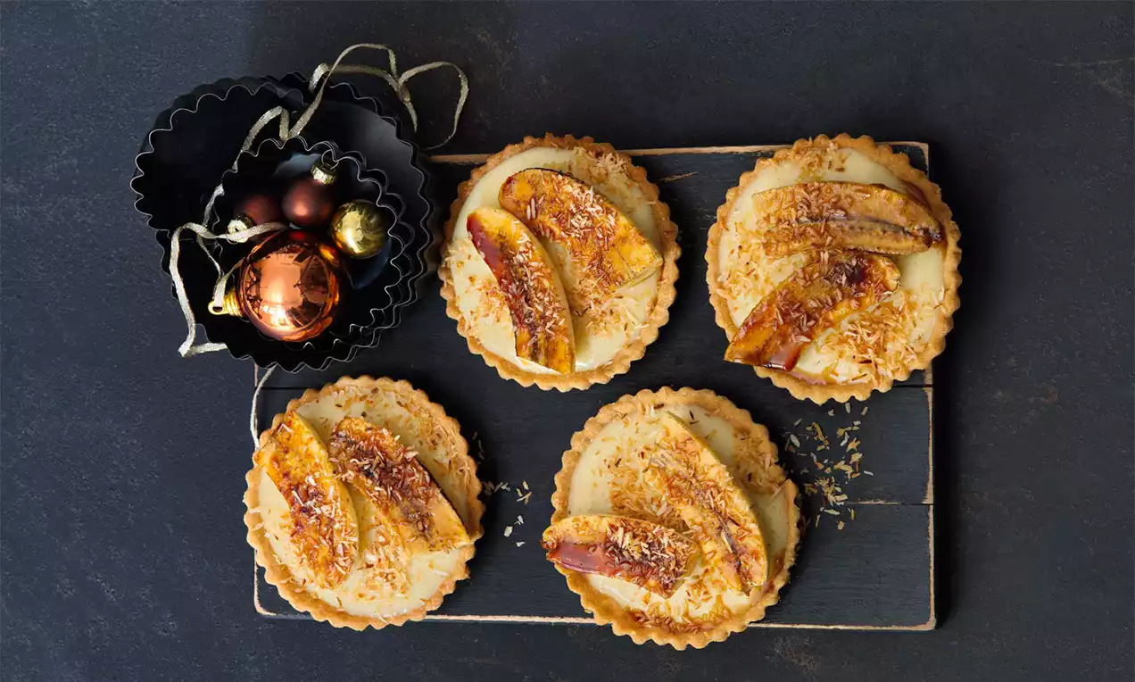 Banánové tartaletky s křehkým světlým těstem, krémovou medovou náplní, opečenými plátky banánu a kokosovými vločkami na tmavém podnosu