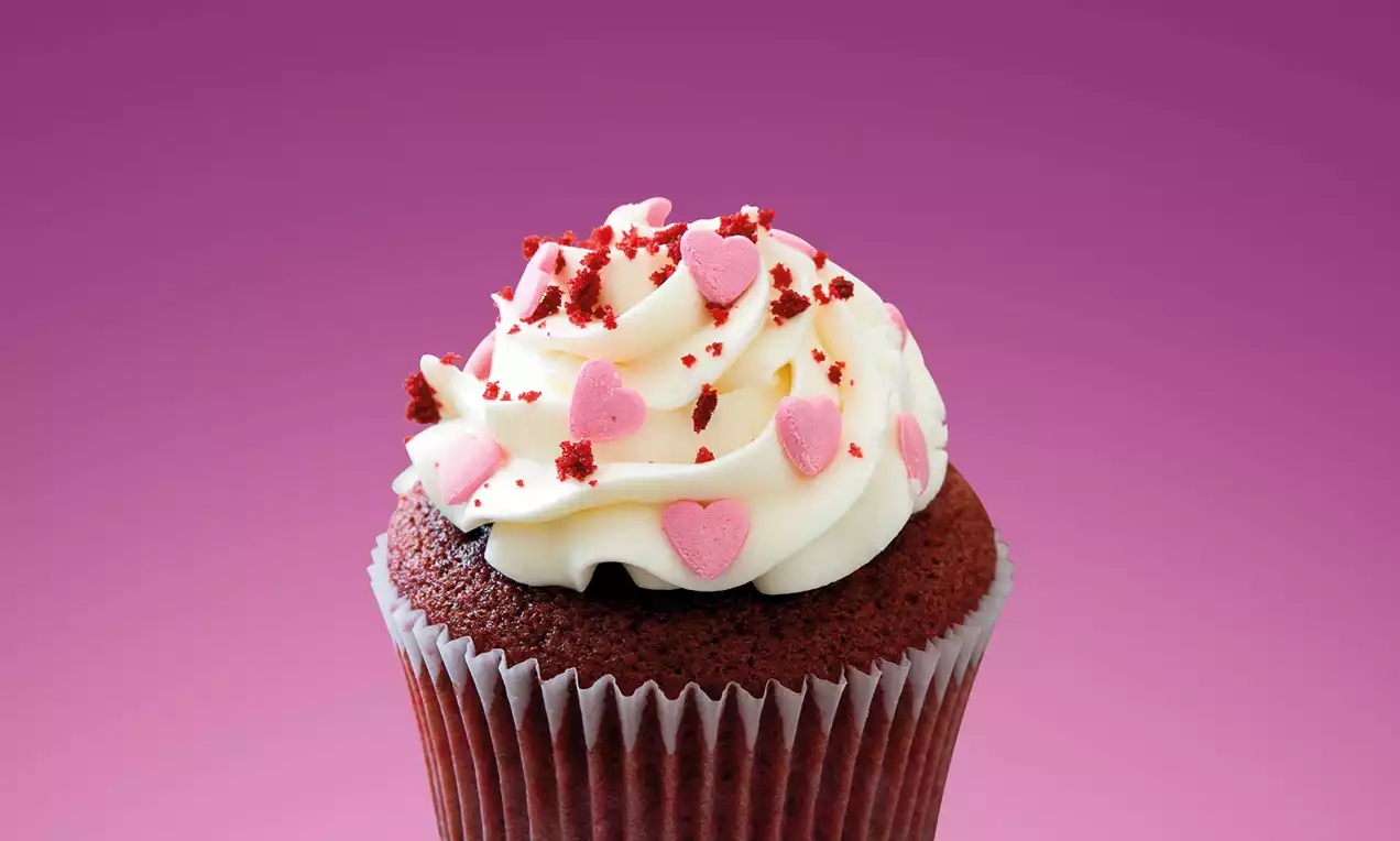 Brioșă pufoasă cu ciocolată în nuanțe de maro închis, decorată cu cremă albă catifelată, inimioare roz și firimituri roșii pe fundal violet