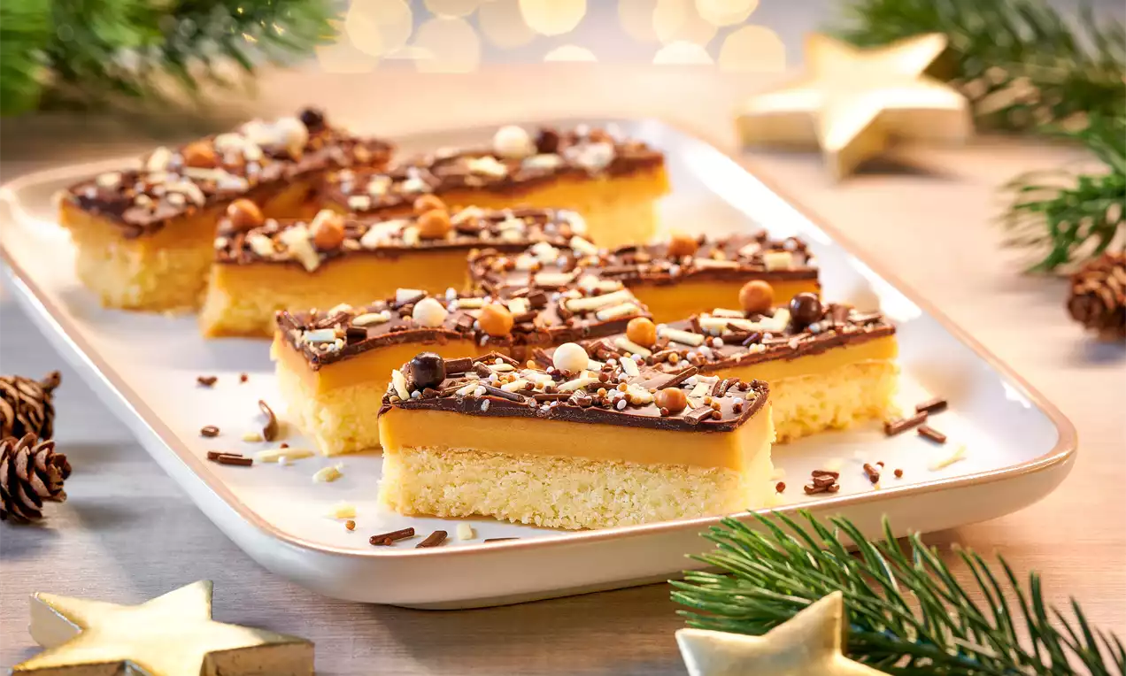 Rechteckige Schoko-Karamell-Shortbreadstücke mit goldener Karamellschicht und Schokoladentopping, garniert mit Streuseln und Crisp-Perlen, auf weißer Platte mit Tannendeko